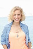 A blonde woman by the sea wearing and apricots top and a denim shirt