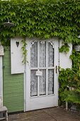 Tür mit Spitzenvorhang hinter Glasfüllung eines Holzhäuschen und berankte Holzfassade