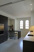 Minimalist kitchen counter opposite fitted cabinets with kitchen appliances and breakfast bar with bar stools; arched windows in background