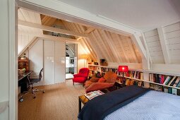 Multifunctional attic room with home office, seating area, bookcases and bed under exposed, white-painted wooden roof structure