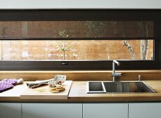 Square, stainless steel sink in kitchen counter with wooden worksurface below ribbon window with translucent roller blind