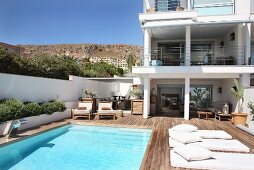 Sonnenbeschienener Pool und Holzterrasse, darauf helle Liegepolster, vor zeitgenössischem Wohnhaus, im Hintergrund mediterrane Landschaft