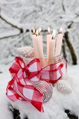 Lit candles in old glass scoop decorated with red ribbon and white Christmas baubles surrounded by snow