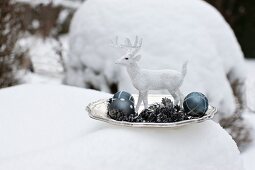 White stag ornament, metal fir cones and Christmas tree baubles on plate surrounded by snow