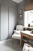 Kitchen table and upholstered chair below window next to grey-painted cupboard door