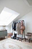 Antique chest of drawers, women's clothing on clothes rail and upholstered chair in bright attic room