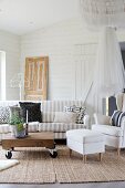White armchair and footstool and striped sofa around wooden table on castors in living room with white wood cladding