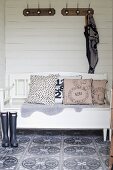 Scatter cushions on white-painted wooden bench below coat racks on white wooden wall and retro-patterned wall tiles