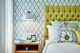 Wooden bedside cabinet below pendant lamp next to bed with yellow headboard against retro wallpaper