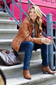 A young woman wearing jeans and a leather jacket sitting on a flight of steps holding a coffee cup