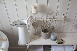 Enamel jug, candlestick and flowering branches in ceramic beaker on old wooden bench in shabby-chic bathroom