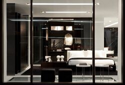 View of bedroom through glass wall; black pouffes, bed with white bed linen and dressing room