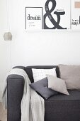 Anthrazitfarbene Couch mit verschiedenen Kissen und Plaid unter weißem Wandboard mit aufgestellten Druckgrafiken