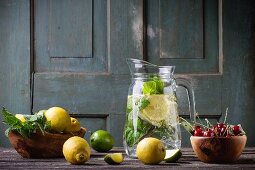 A glass jug of homemade lemonade with lemons, limes, mint and cherries on a wooden table