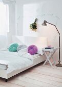 Modern double bed with white headboard and round, colourful scatter cushions; folding bedside table and retro standard lamp