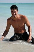 A young, topless man wearing bathing short keeling on the beach