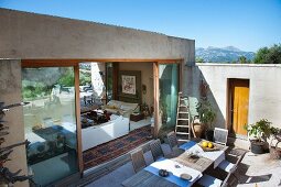 Large wooden dining table on terrace of Mediterranean bungalow with view into interior