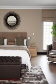 Dark brown ottoman at foot of double bed and sunburst mirror on beige-painted wall