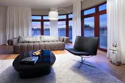 Eleganter Sitzbereich mit Ledersessel und Hochglanztisch vor geschwungener Fensterfront in der Abenddämmerung
