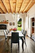 Long dining table with postmodern curved legs below designer lamp with glass plate hanging from wood-beamed ceiling in spacious kitchen