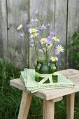 Delicate campanula and ox-eye daisies in green glass bottles on folded green and white tablecloth on rustic wooden stool