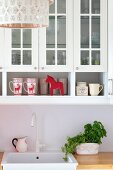 Mugs with reindeer motif below glass-fronted wall units above white sink and potted herbs