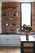 Open-fronted, suspended shelves against brick wall above white base units and blue island counter in open-plan kitchen