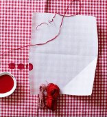 Weißes Blatt Papier auf rot-weiß kariertem Stoff mit verschiedenen Garnrollen und roter Farbe