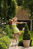 A woman dancing in a garden wearing a light-green slip with a wicker bag