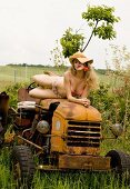 A woman wearing underwear and a straw hat lying on an old tractor with a flower in her mouth