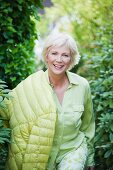 An older woman wearing a light green blouse, green-and-white trousers and a quilted jacket in a garden