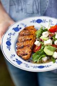 A woman holding a plate of grilled pork collar steak and rocket salad with tomatoes, cucumber and feta cheese