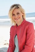 A blonde woman by the sea wearing a light jumper and a salmon pink leather jacket