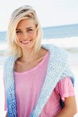 Blonde Frau mit rosa Shirt und Pulli über Schultern am Strand