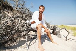 A young man wearing an open shirt and shorts sitting on a beach on driftwood