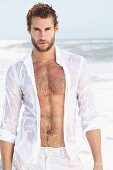 Junger Mann in nassem, weissen Hemd und Bermudashorts steht am Meer