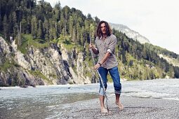 A young man with a beard and long hair walking along a gravelly river bed