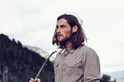 A young man with a beard and long hair against a wooded mountain