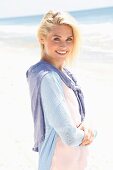 Lächelnde blonde Frau in pastellfarbenem Shirt, Jacke und Hose steht am Strand