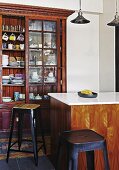 Retro bar stools around counter in front of crockery in antique glass-fronted cabinet