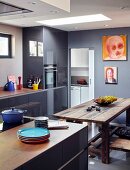 Stack of colourful plates on free-standing counter next to rustic dining set in modern kitchen with artworks on grey-blue wall