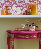 Pink, ornate console table under white floating shelf and floral wallpaper