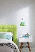 A modern bedside table next to a bed with a lime green padded headboard against a wall with a light-coloured liberty pattern