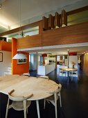 View across dining table into open-plan, designer kitchen below wood-clad gallery with orange partition wall