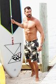 A dark-haired man on a beach wearing bathing shorts with a surfboard