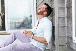 Dunkelhaariger Mann mit Bart in weißem Hemd und Karohose sitzt lachend auf dem Boden
