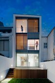 View from garden to wooden terrace and contemporary three-storey house with illuminated interior at twilight