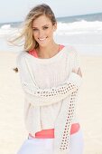 A young woman on a beach wearing a pink top and white knitted jumper
