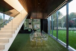 Transparent dining set on artificial lawn between staircase in contemporary house and glass wall with view of garden