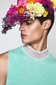 A young man wearing a turquoise sleeveless jumper with flowers on his head